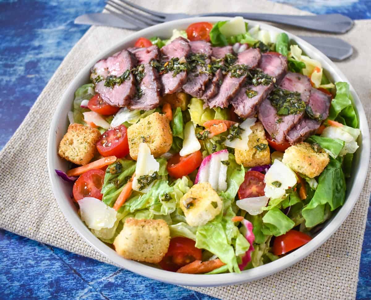 The chimichurri steak salad in a white bowl and set on a beige linen on a blue table.