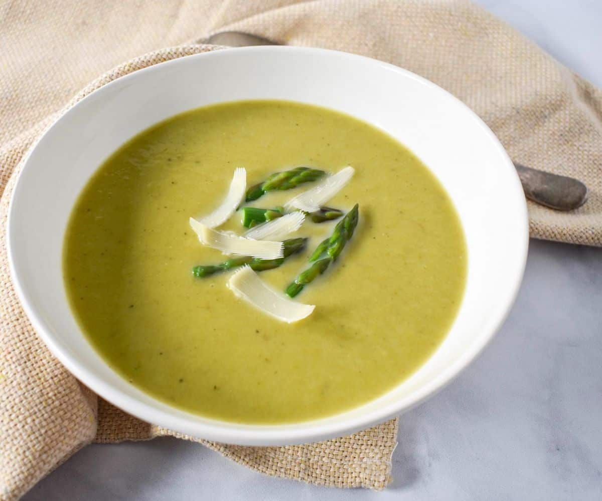 The cream of asparagus soup garnished with asparagus tips and parmesan cheese and served in a white bowl with a beige linen and a spoon.