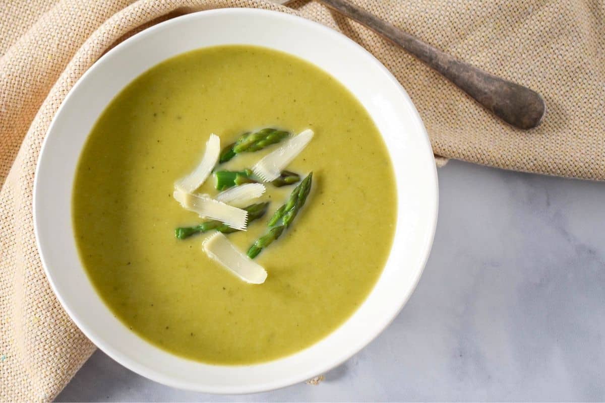 The asparagus soup garnished with asparagus tips and parmesan cheese and served in a white bowl with a beige linen and a spoon.
