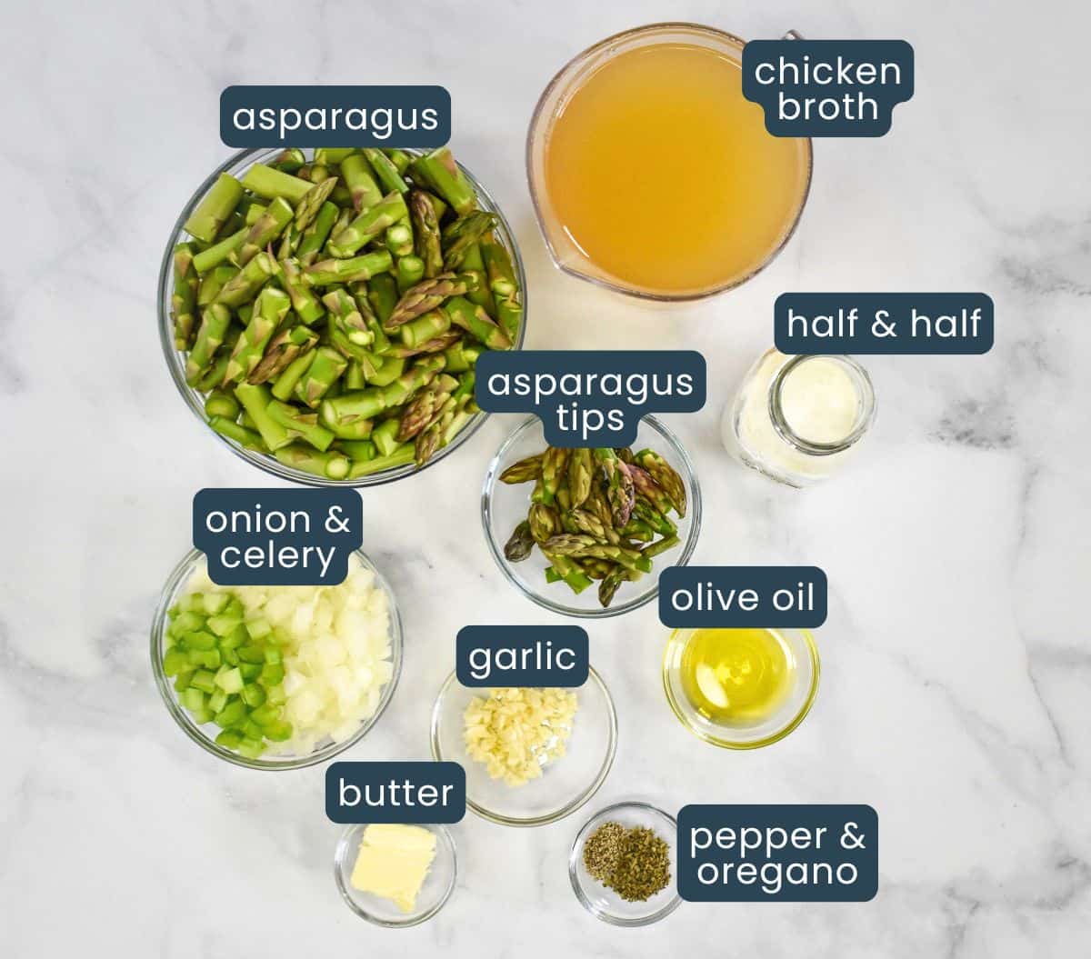 The ingredients for the cream of asparagus soup prepped and separated in glass bowls arranged on a white table, with each labeled.