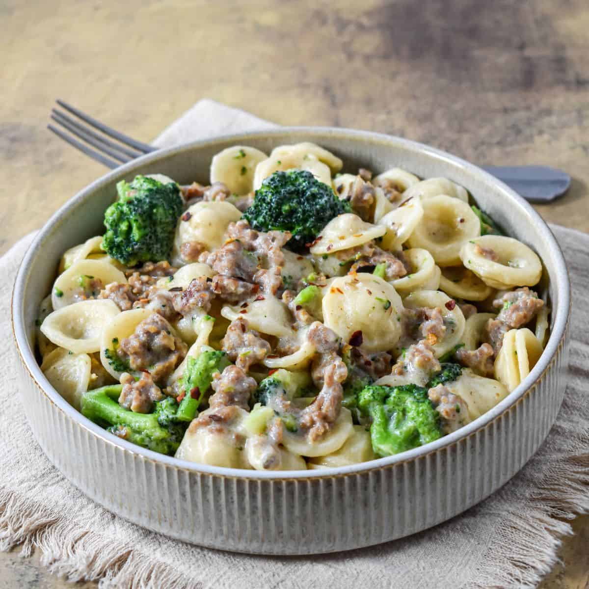 Bowl of creamy orecchiette with sausage and broccoli served on a beige napkin with a fork in the background.