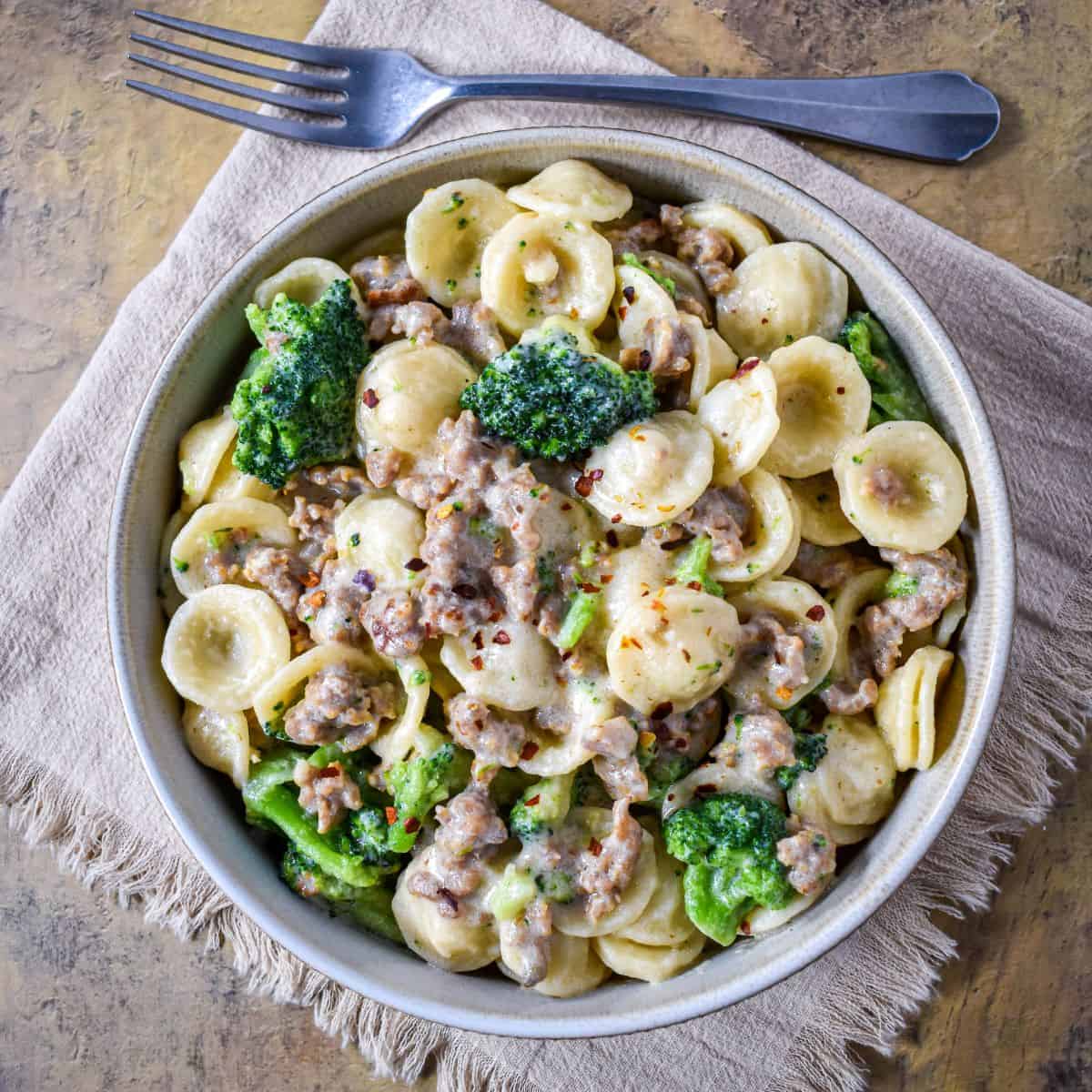 Top view of creamy orecchiette with sausage and broccoli in a light gray bowl on a textured background.