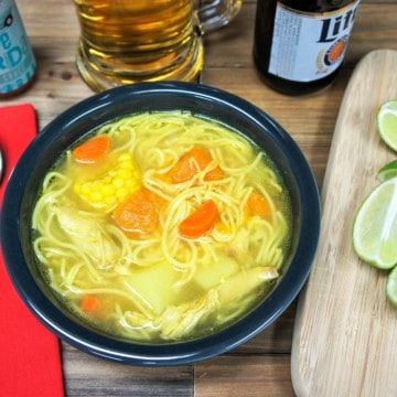Cuban chicken soup with noodles carrots, potatoes, pumpkin, corn and chicken served in a dark gray bowl with lime wedges and a beer mug in the background.