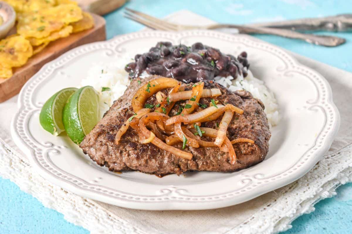 Close-up of Cuban cube steak with sautéed onions, rice, black beans, and lime on a white plate.