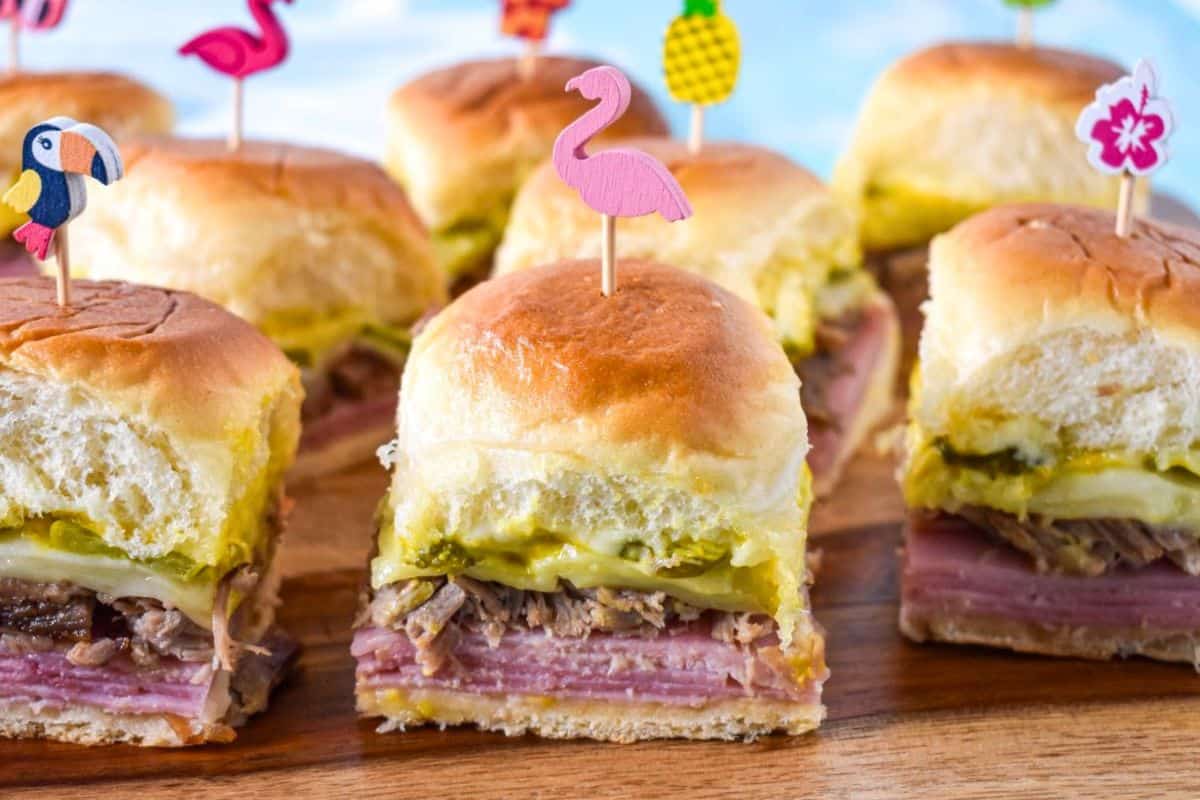 Cuban sliders arranged on a wood cutting board, filled with roast pork, ham, Swiss cheese, pickles, and topped with decorative toothpicks.