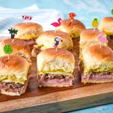 Cuban sliders arranged on a wood cutting board, each slider topped with colorful tropical-themed toothpicks.