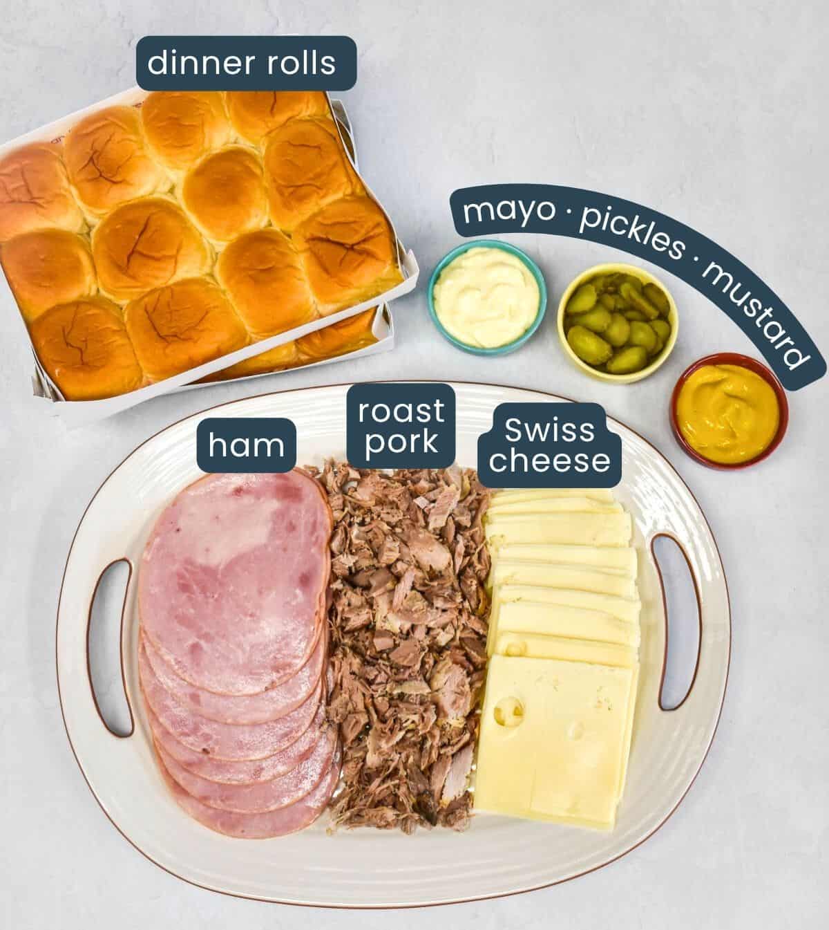 Ingredients for Cuban sliders displayed on a platter and labeled: ham, roast pork, Swiss cheese, dinner rolls, mayonnaise, pickles, and mustard.
