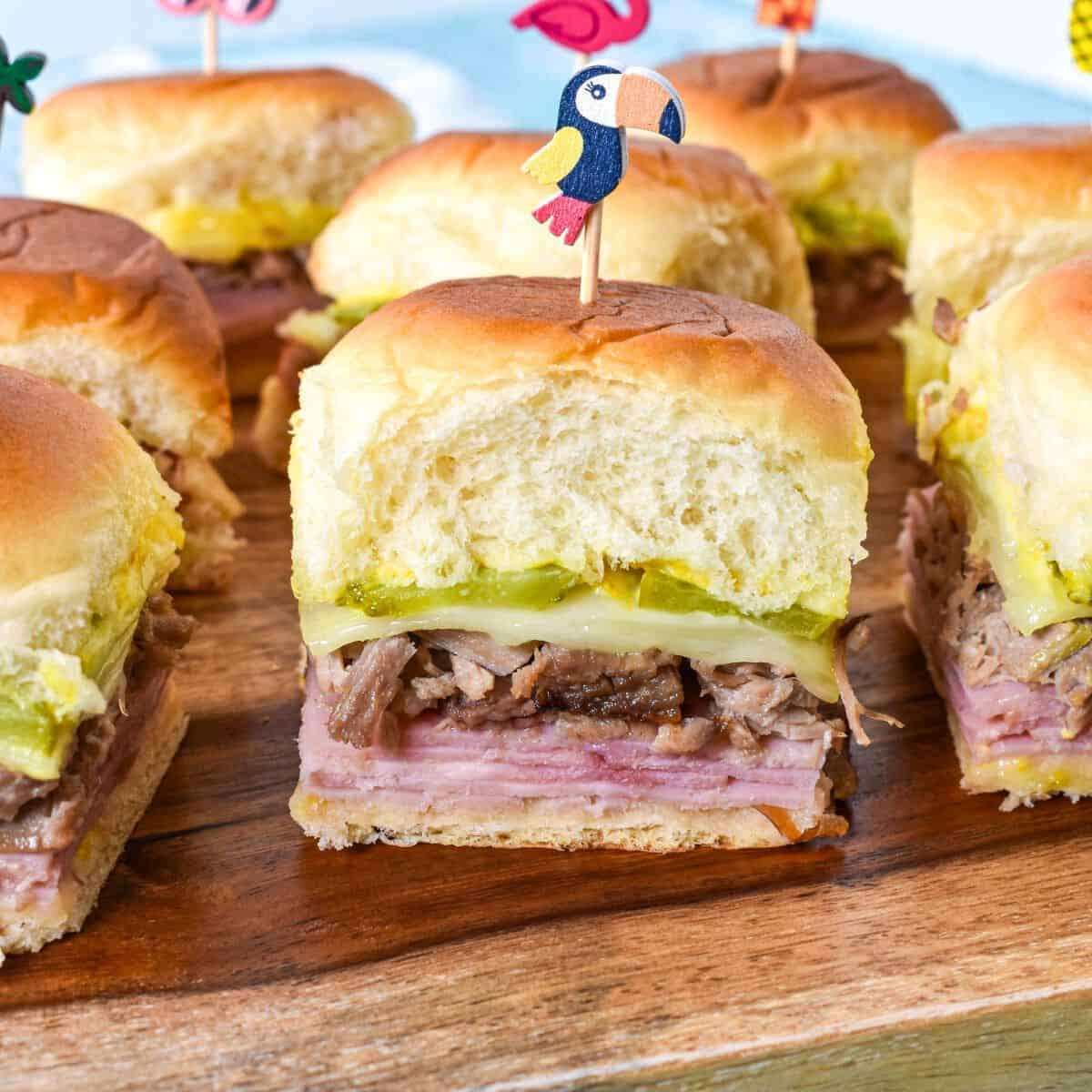 Close-up of a Cuban slider with visible layers of ham, roast pork, pickles, and Swiss cheese inside a soft roll, topped with a toucan-themed toothpick.