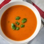 An image of the tomato soup served in a white bowl on a white linen with a red stripe and a spoon to the top, right side.