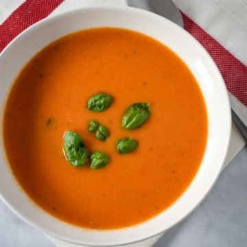 An image of the tomato soup served in a white bowl on a white linen with a red stripe and a spoon to the top, right side.