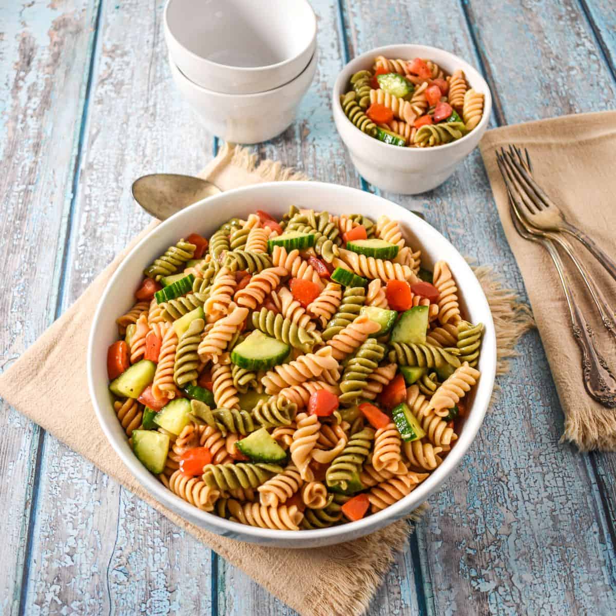 Garden pasta salad in a large white serving bowl with a smaller portion, stacked bowls, and utensils on a blue rustic table.