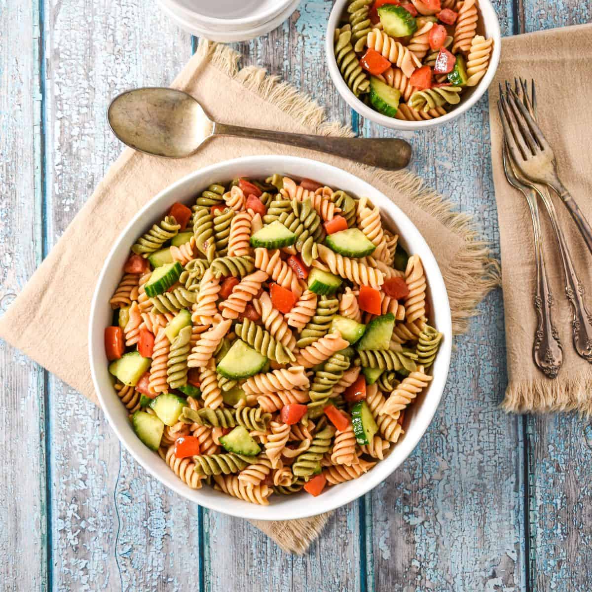 Garden pasta salad in a large white serving bowl with a smaller portion, stacked bowls, and utensils on a blue rustic table.