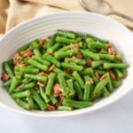 An image of the green beans with bacon served in an oval white bowl with a beige linen and a serving spoon to the right.