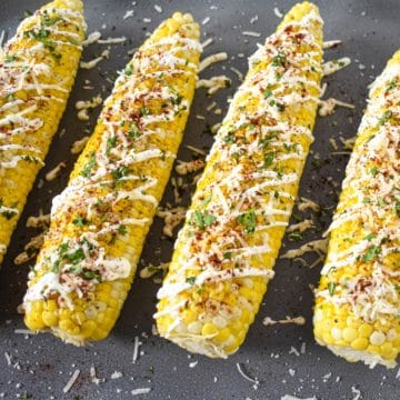 Four corn cobs topped with sour cream, chili powder, parmesan, and cilantro. They are set on a gray platter.