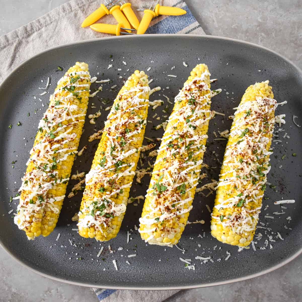 Four corn cobs topped with sour cream, chili powder, parmesan, and cilantro. They are set on a gray platter with yellow corn holders on top.