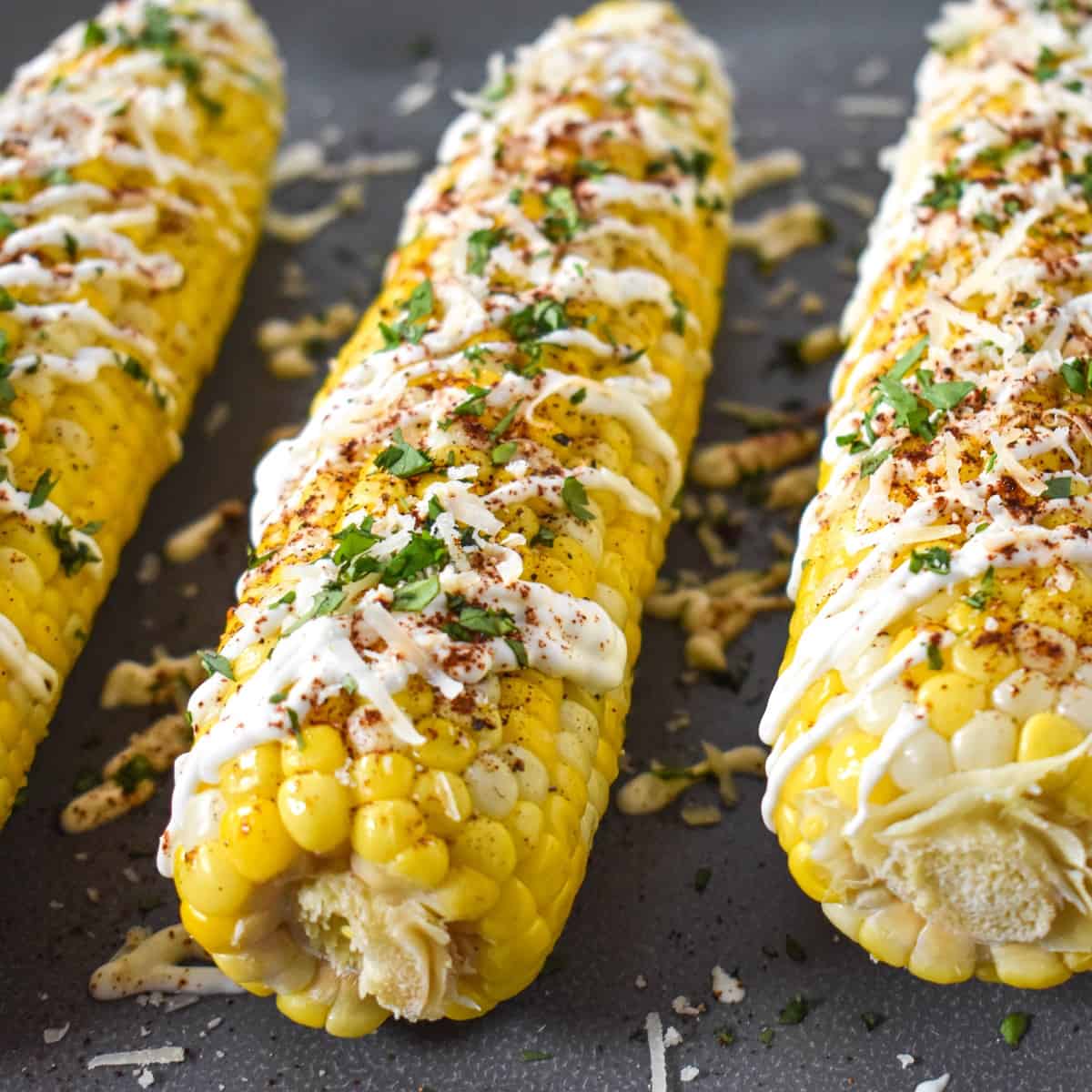 A close-up of the cobs topped with sour cream, chili powder, parmesan, and cilantro, set on a gray platter.