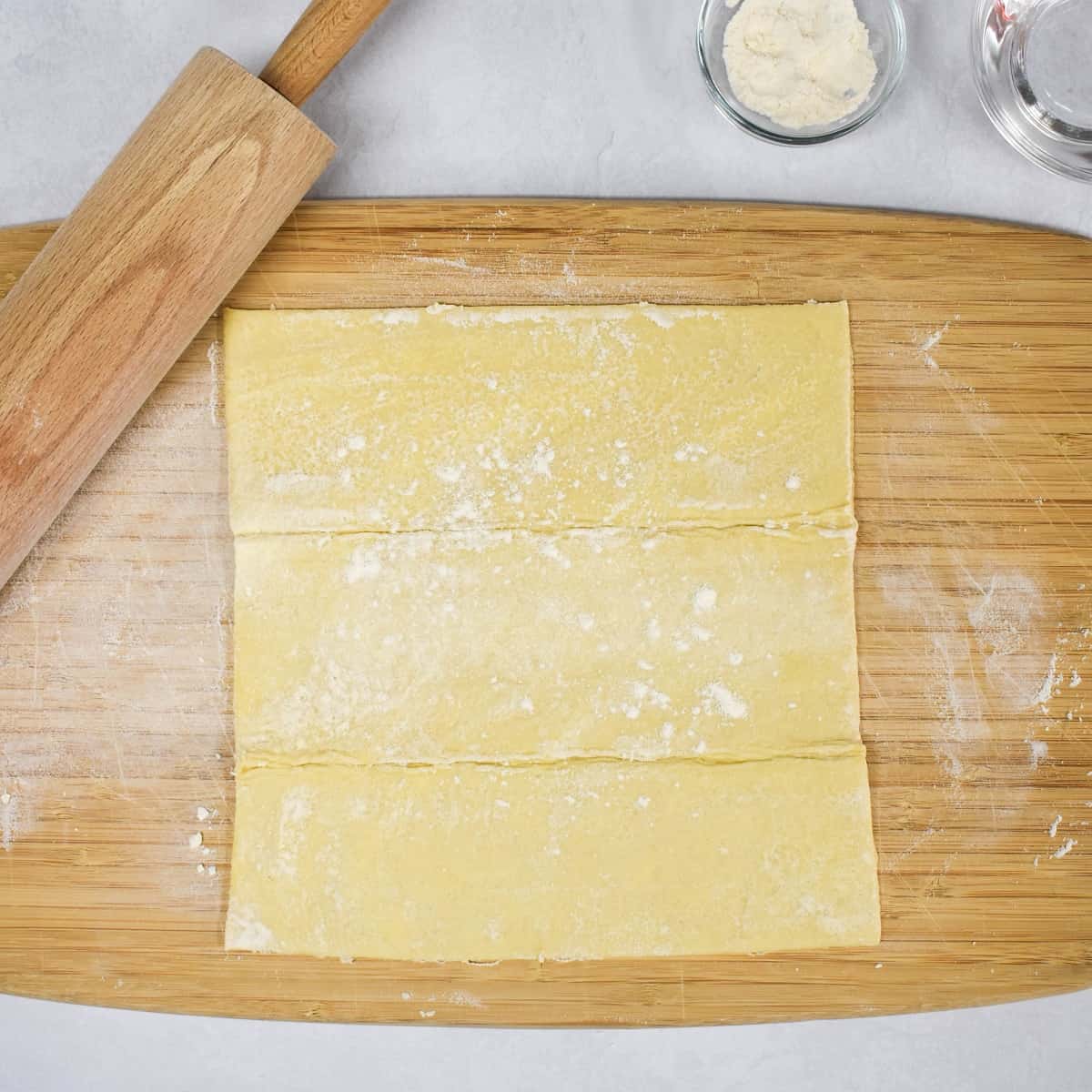 A puff pastry sheet on a floured wood cutting board with a rolling pin on the side.
