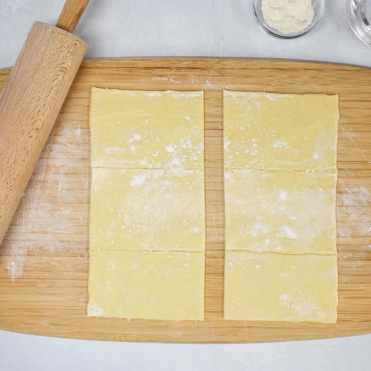 A puff pastry sheet cut in half on a wood cutting board with a rolling pin to the side.
