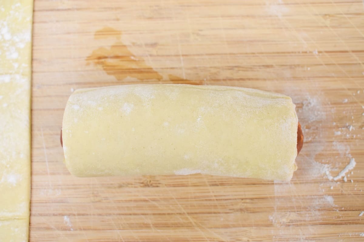 The rolled puff pastry covering the sausage set on a wood cutting board.