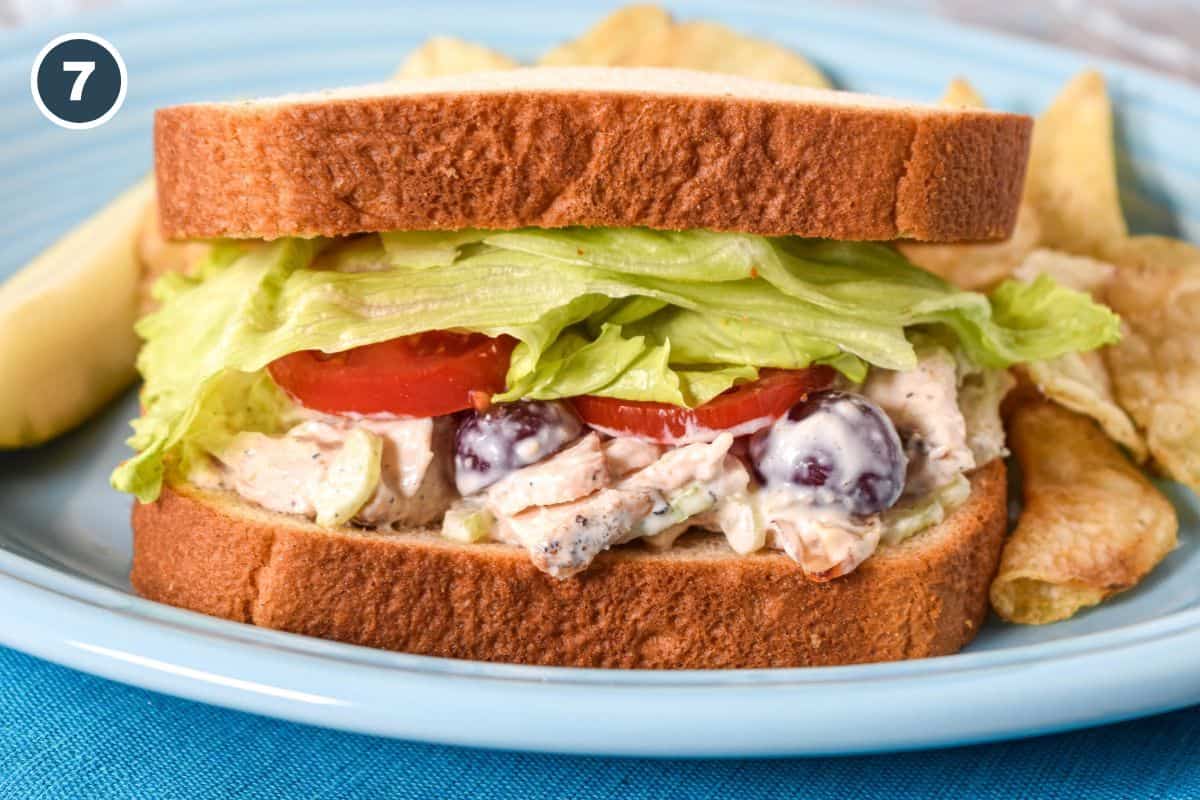 Chicken salad sandwich with lettuce and sliced tomatoes, served with potato chips and a pickle.