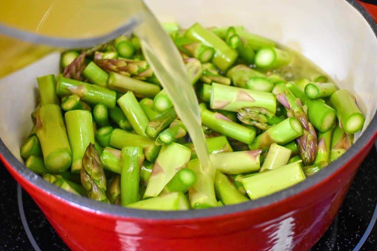 Chicken broth being poured over fresh asparagus and vegetables in a large pot.