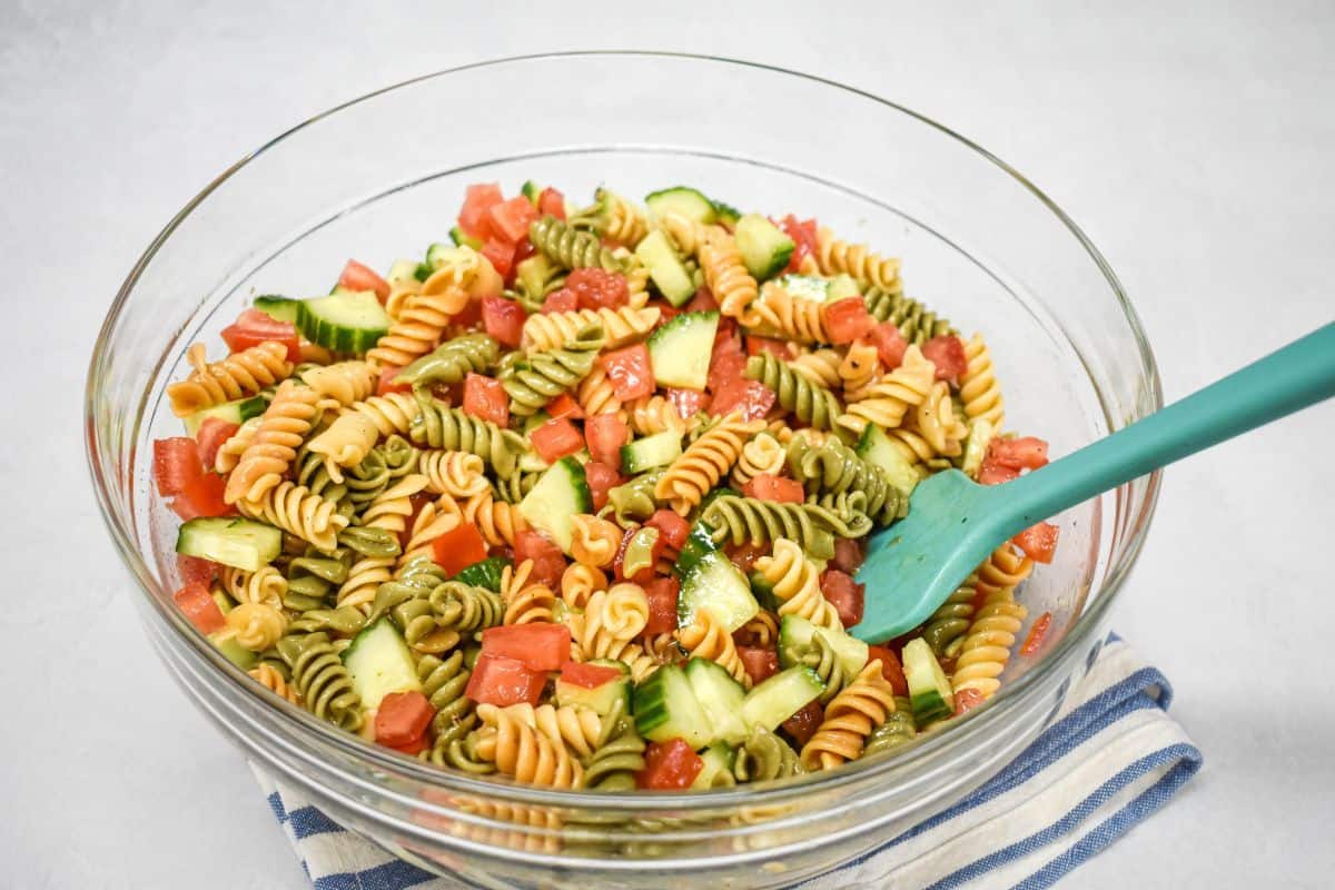 Garden pasta salad with tomatoes and cucumber being mixed in a glass bowl.