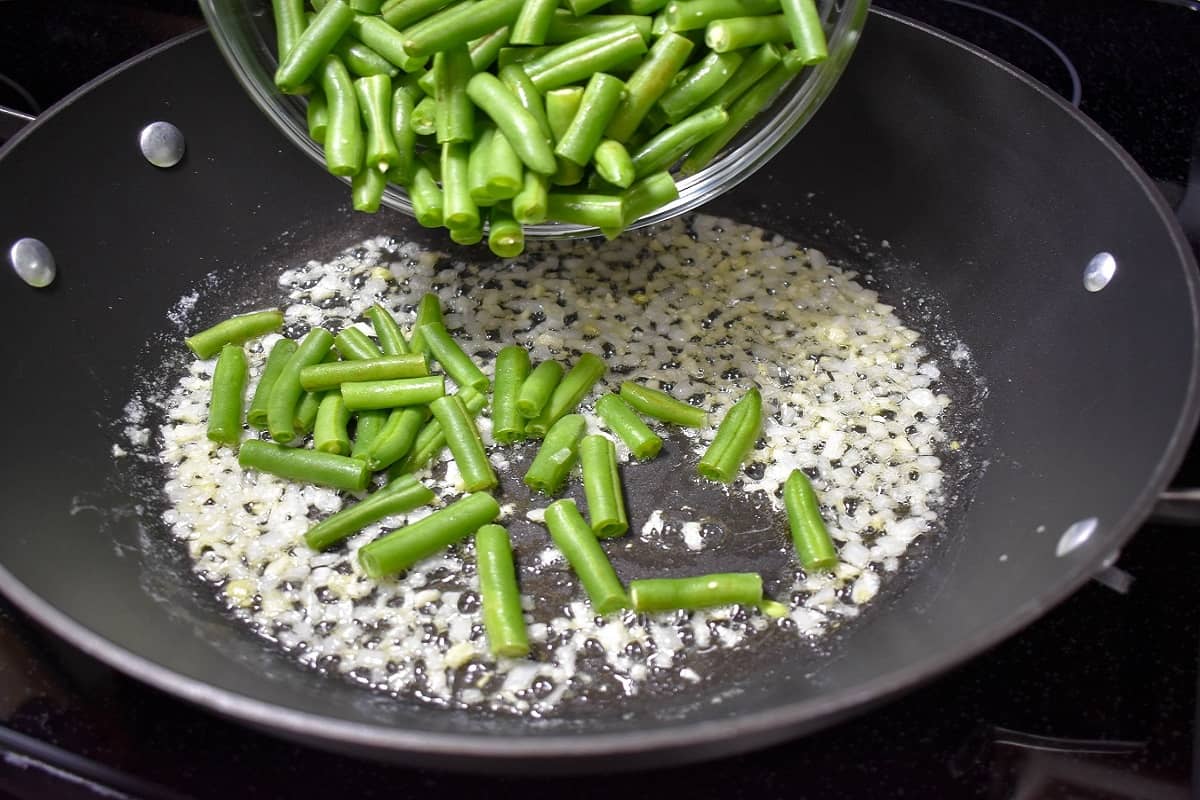 Green beans being added to onions and garlic cooking in a large, black skillet.