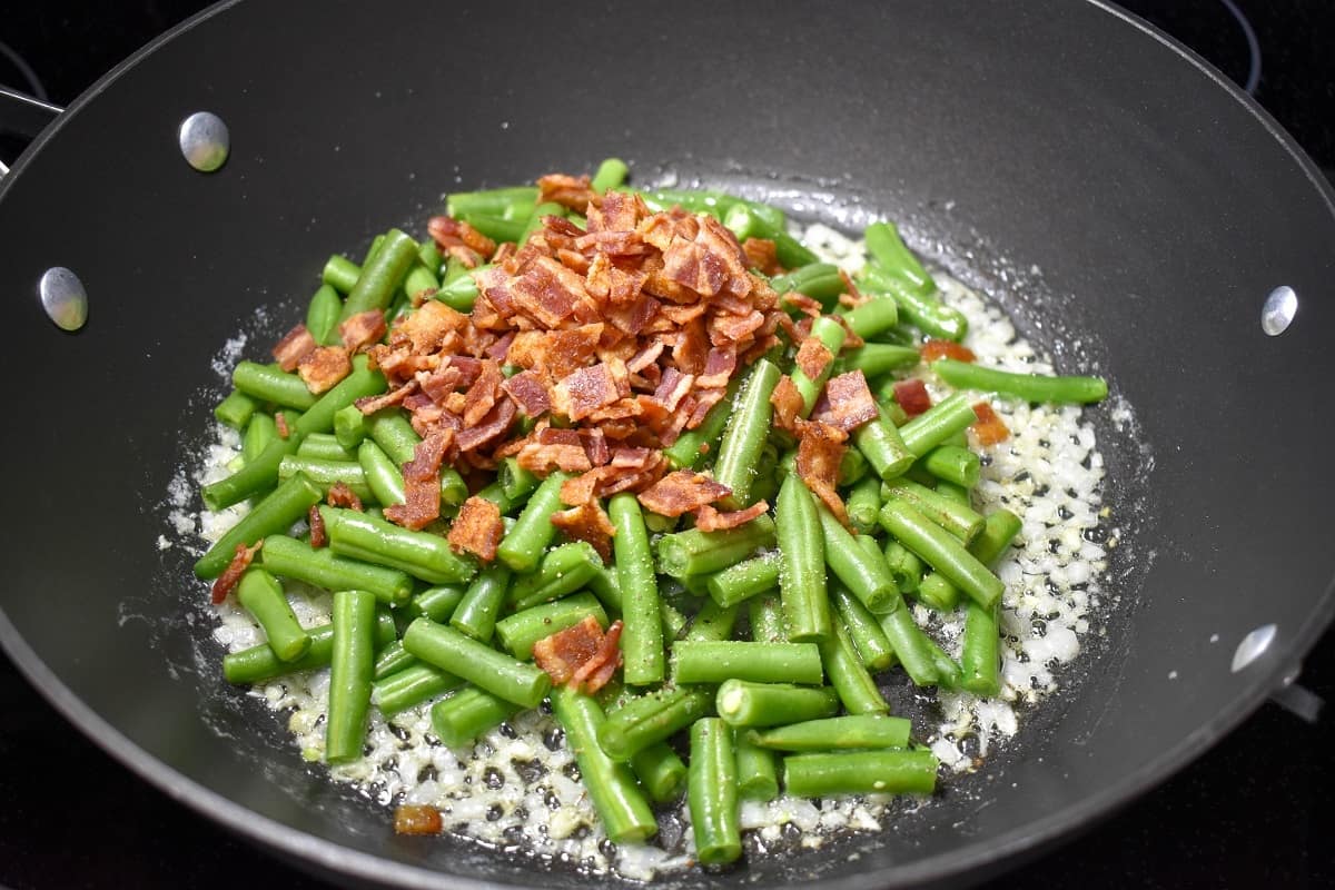 Crumbled bacon added to the top of the green beans and onions cooking in a large, black skillet.