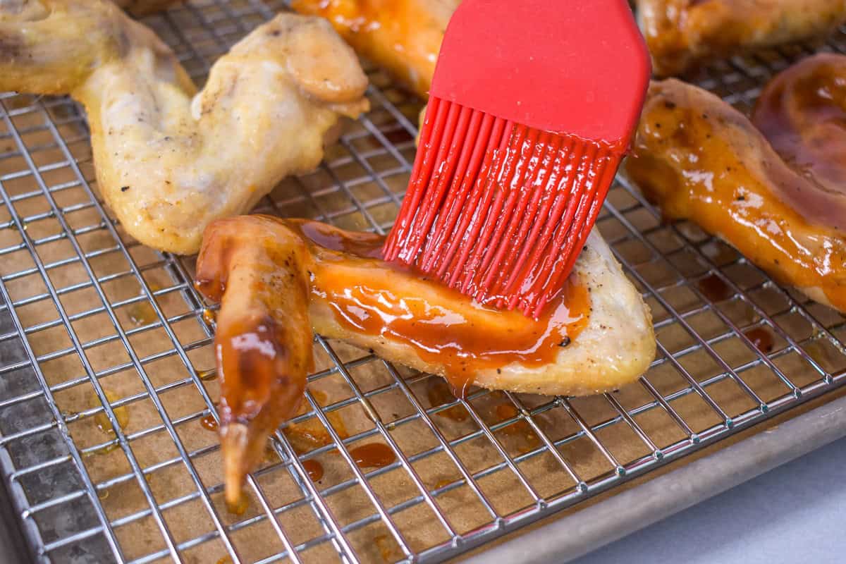 Wings being brushed with sauce with a red silicone pastry brush on a sheet pan lined with a cooling rack.