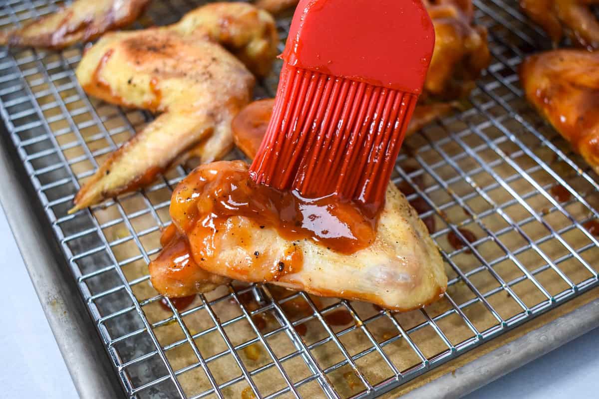 Wings being brushed with sauce with a red silicone pastry brush on a sheet pan lined with a cooling rack.