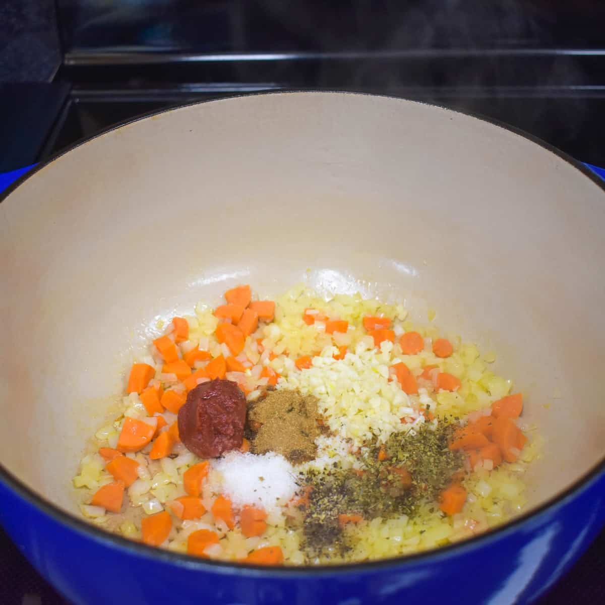 An image of onions, carrots, garlic, tomato paste, and spices in a large blue pot.