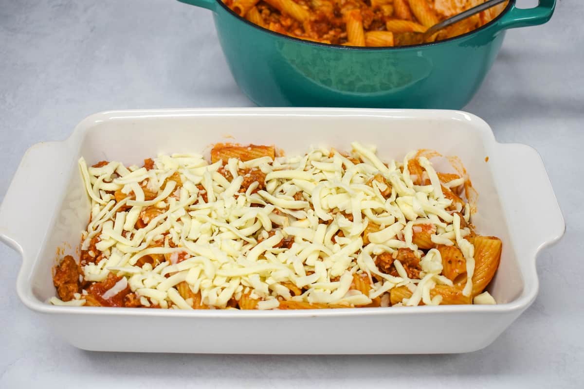 Half of the rigatoni in a white baking dish sprinkled with shredded mozzarella cheese on top.