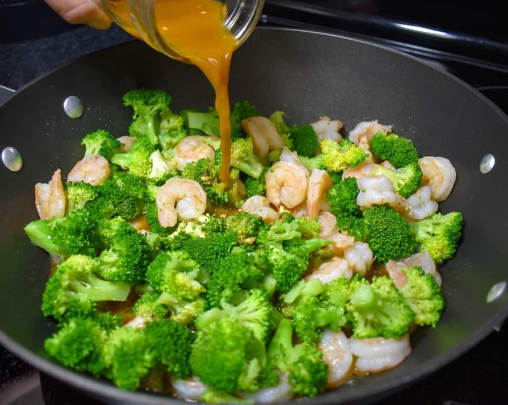 An image of the prepared sauce being added to the shrimp and broccoli in a large, black skillet.