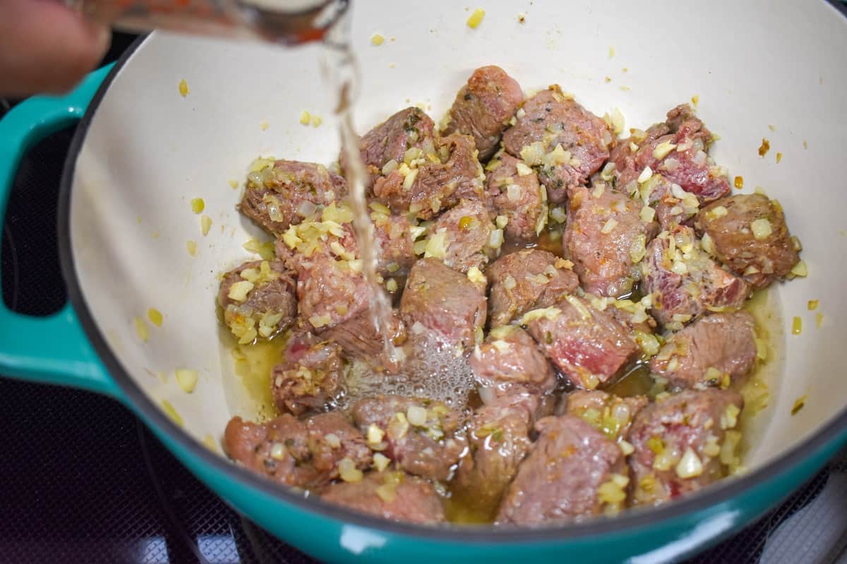 White wine being added to lightly browned beef chunks in a large pot.