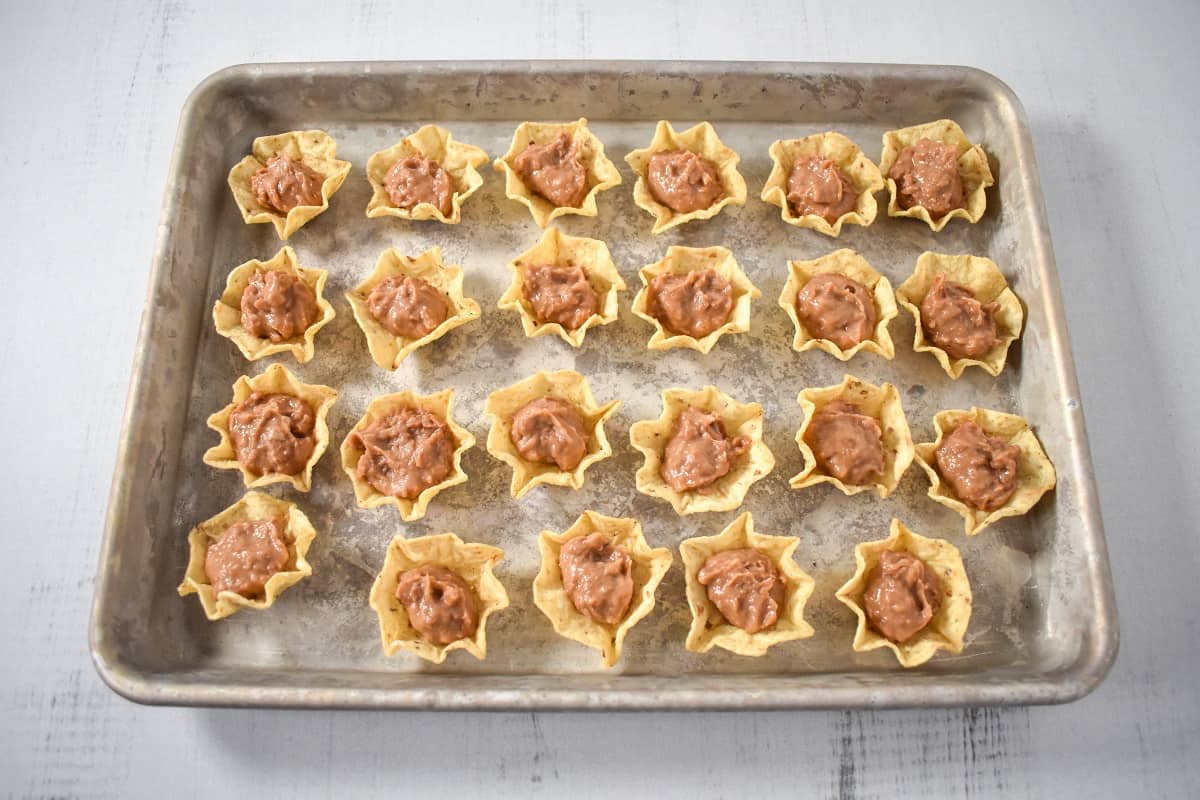 An image of tortilla scoop chips filled with refried beans and arranged on a baking sheet.