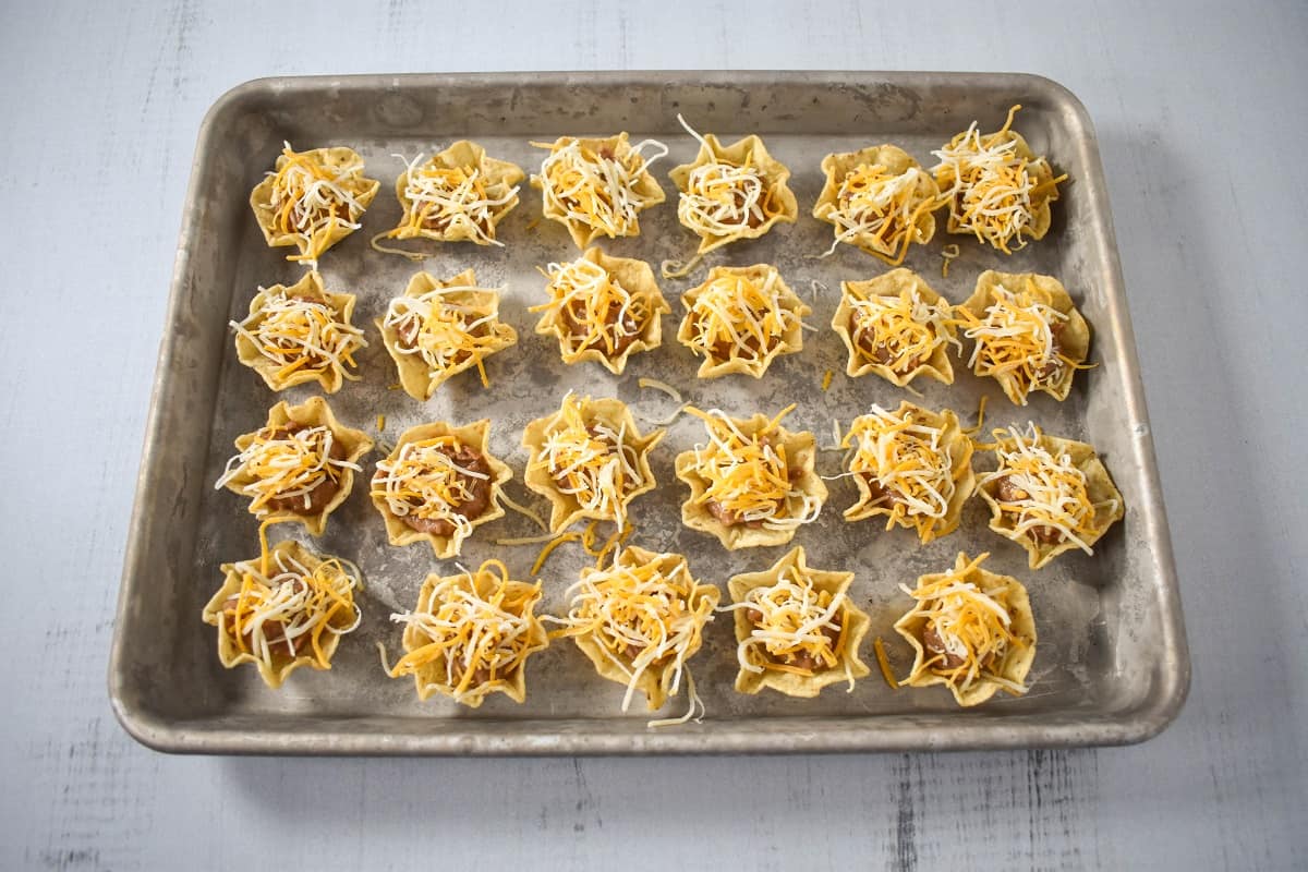 An image of tortilla scoop chips filled with refried beans, topped with a pinch of shredded cheese, and arranged on a baking sheet.