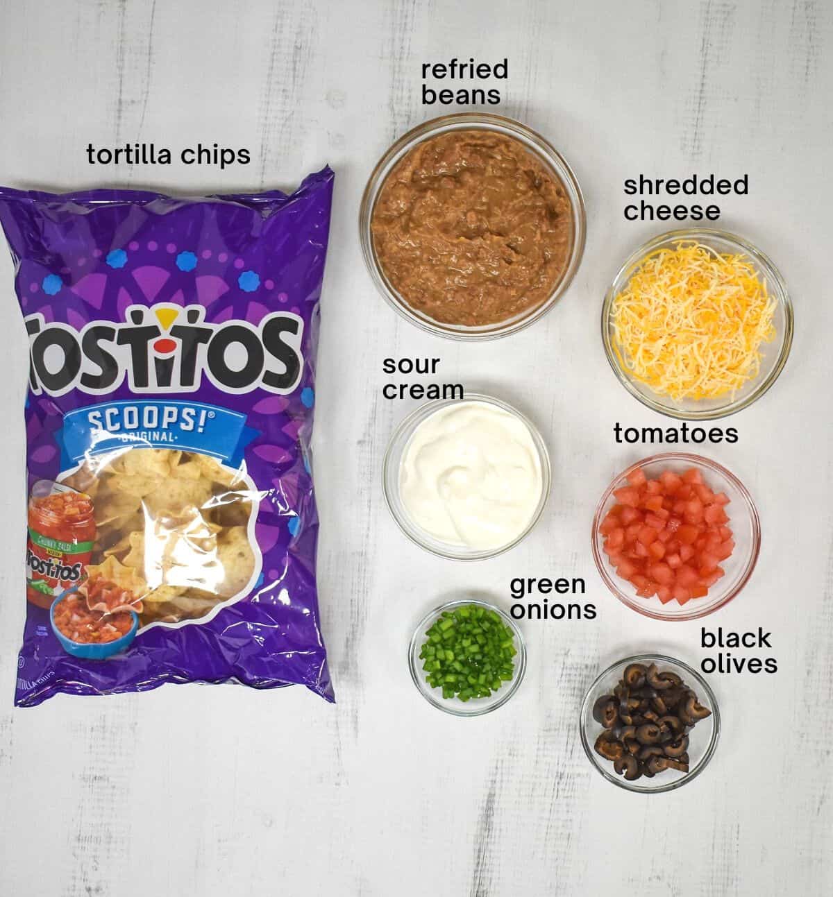 The ingredients for the mini tacos arranged in glass bowls on a white table. The chips are still in the bag.