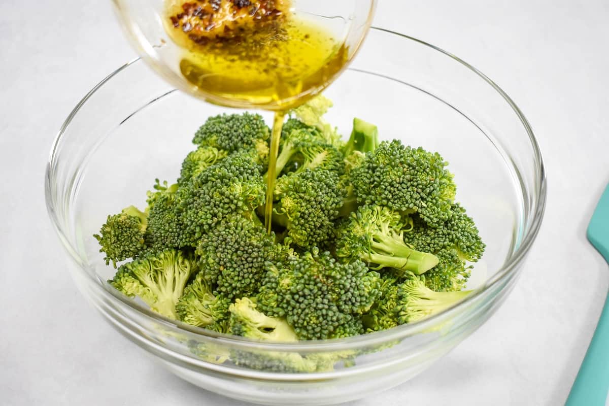 The prepared olive oil being poured on broccoli florets in a large, glass bowl.