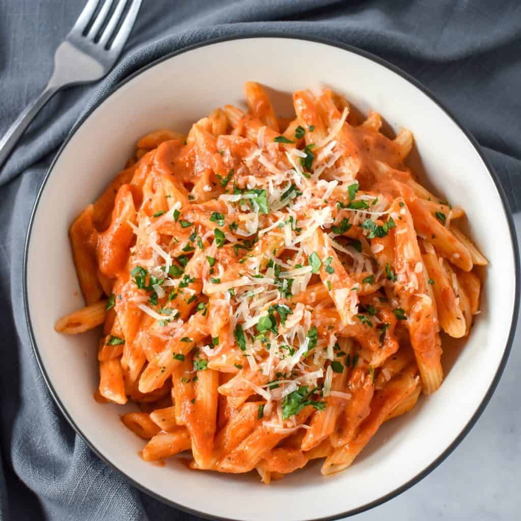 Pasta with vodka sauce garnished with chopped parsley and parmesan cheese, served in a white bowl with a black rim with a gray linen and a fork in the upper right hand side.