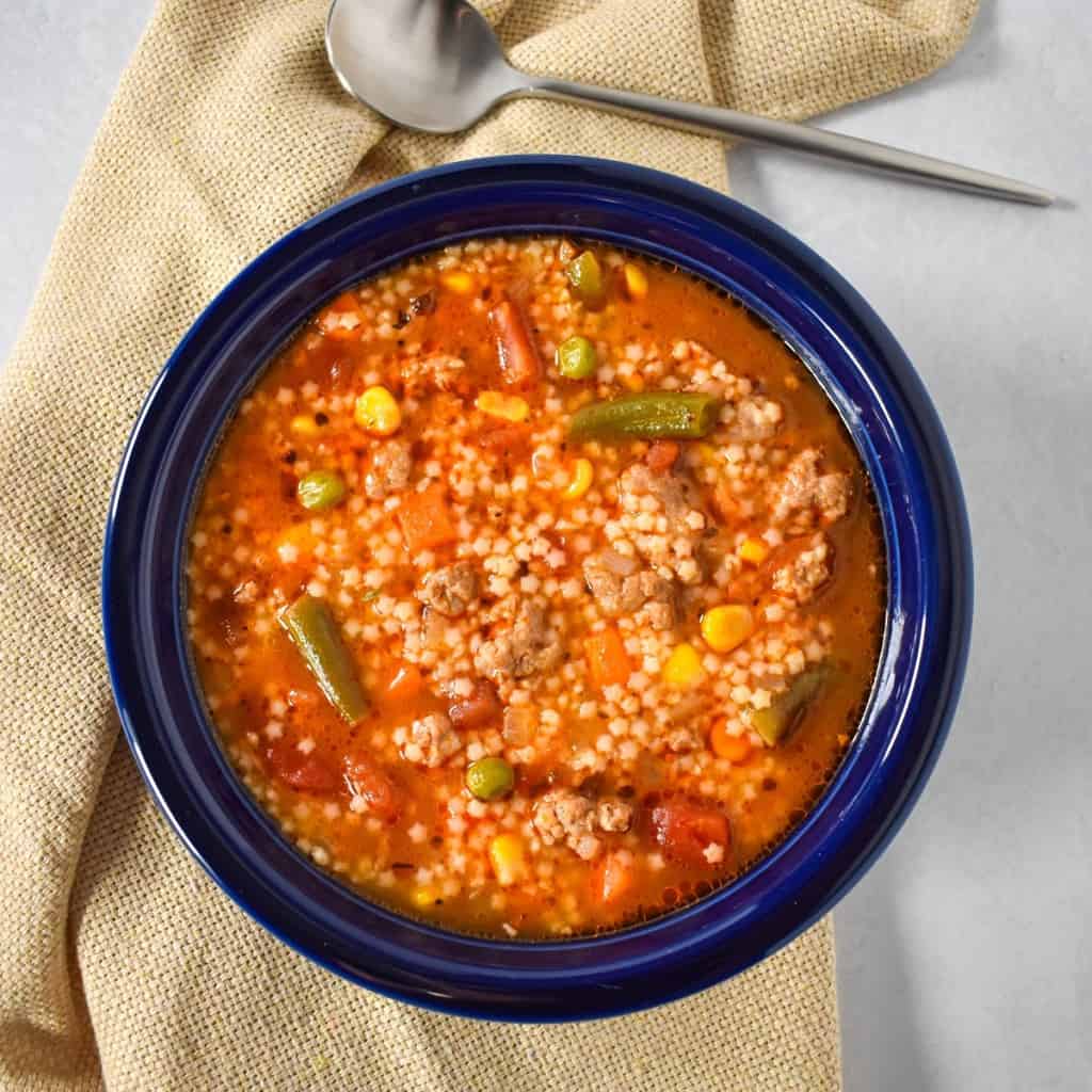 An image of the Italian sausage pastina soup served in a blue bowl with a beige linen set on a white table with a spoon to the top right.