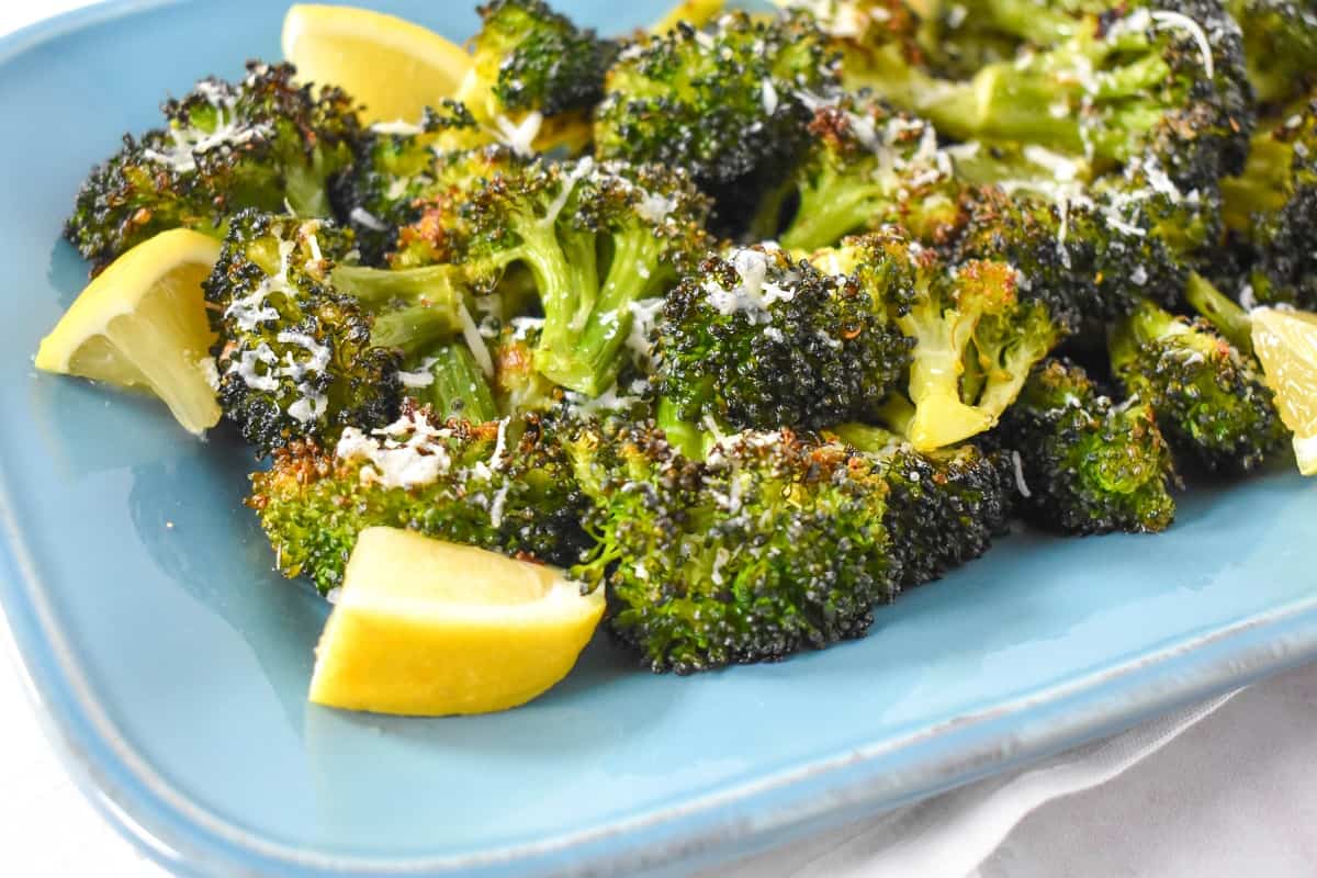 A close up of the finished broccoli garnished with parmesan and lemon wedges served on a light blue plate.