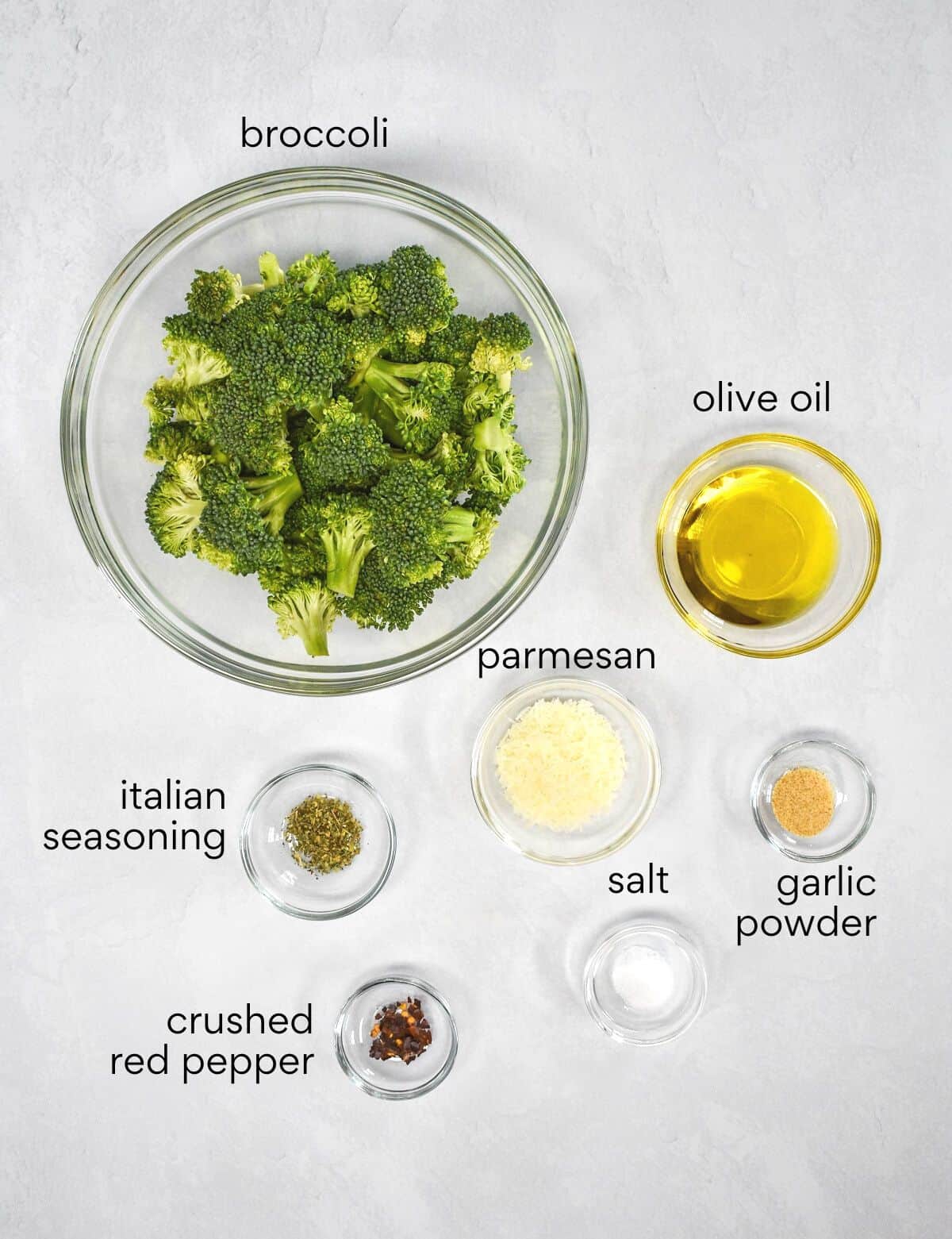 The ingredients for the roasted broccoli prepped and arranged in glass bowls on a white table. Each ingredient is labeled with small black letters.