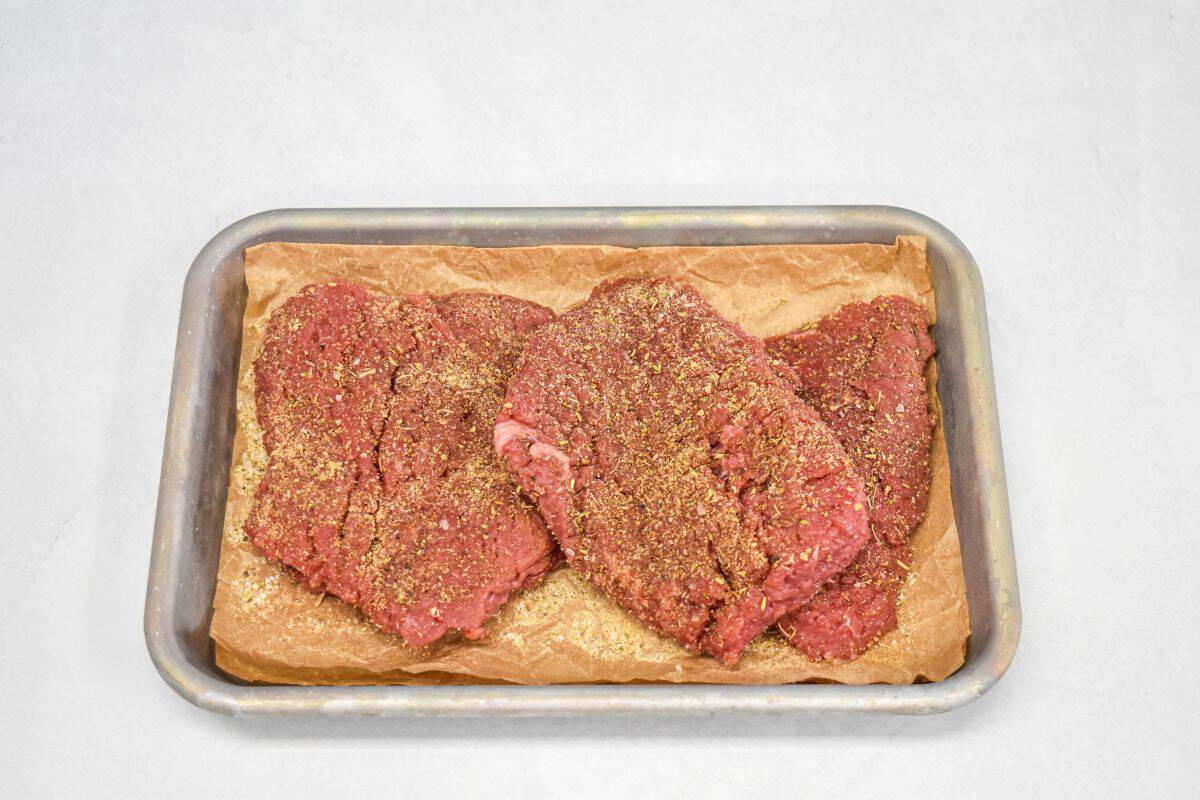 Raw cube steaks seasoned with spices on a baking sheet ready to cook.