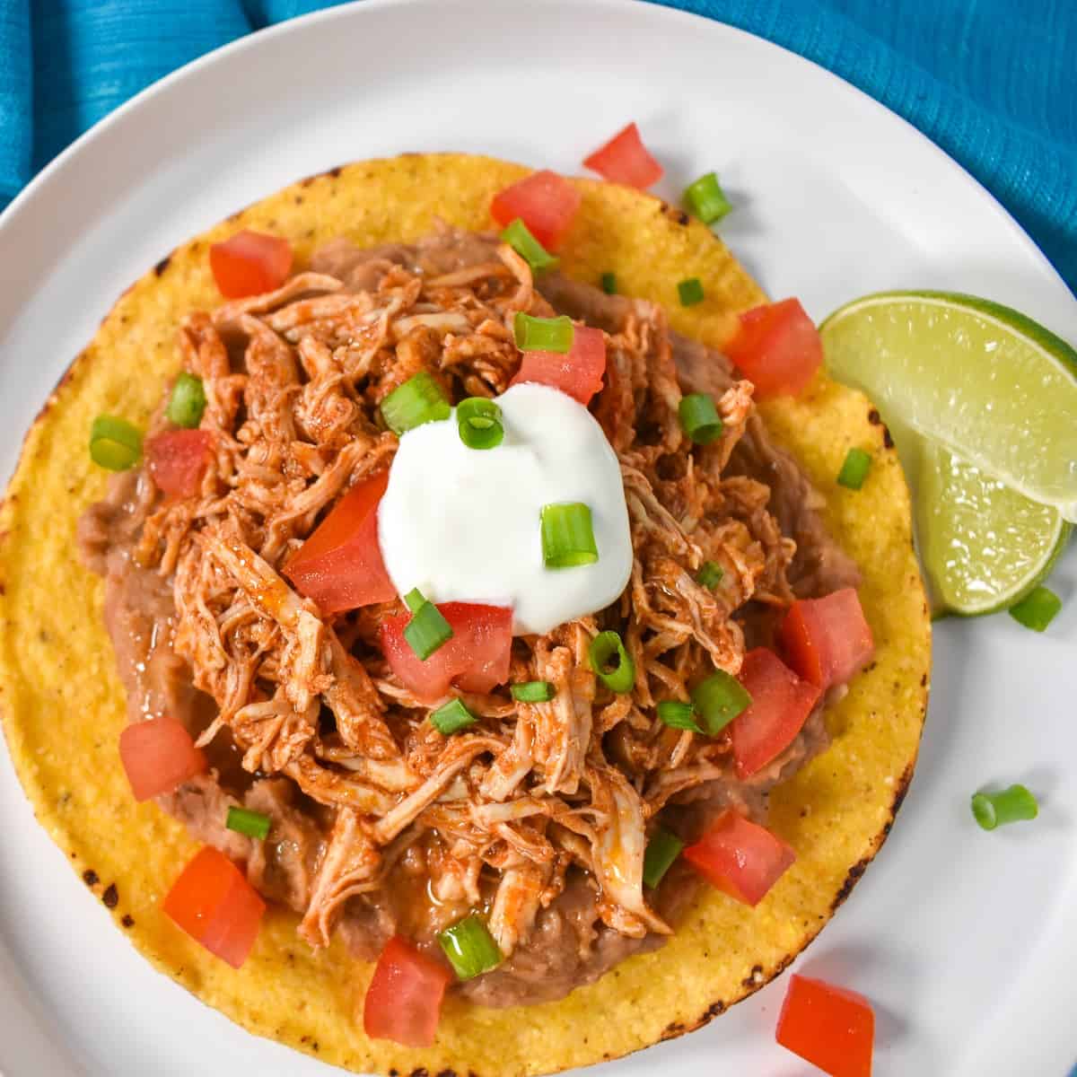 An image of the shredded chicken tostada on a white plate and garnished with diced tomatoes, sour cream and sliced green onions with two lime wedges on the side.