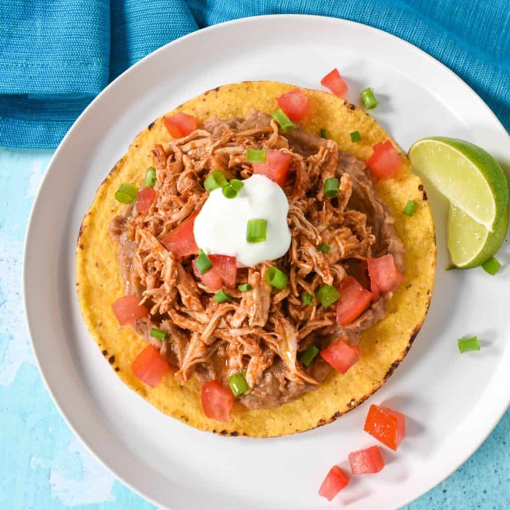 An image of the shredded chicken tostada on a white plate and garnished with diced tomatoes, sour cream and sliced green onions with two lime wedges on the side. The plate is set on a light blue table with an aqua linen.