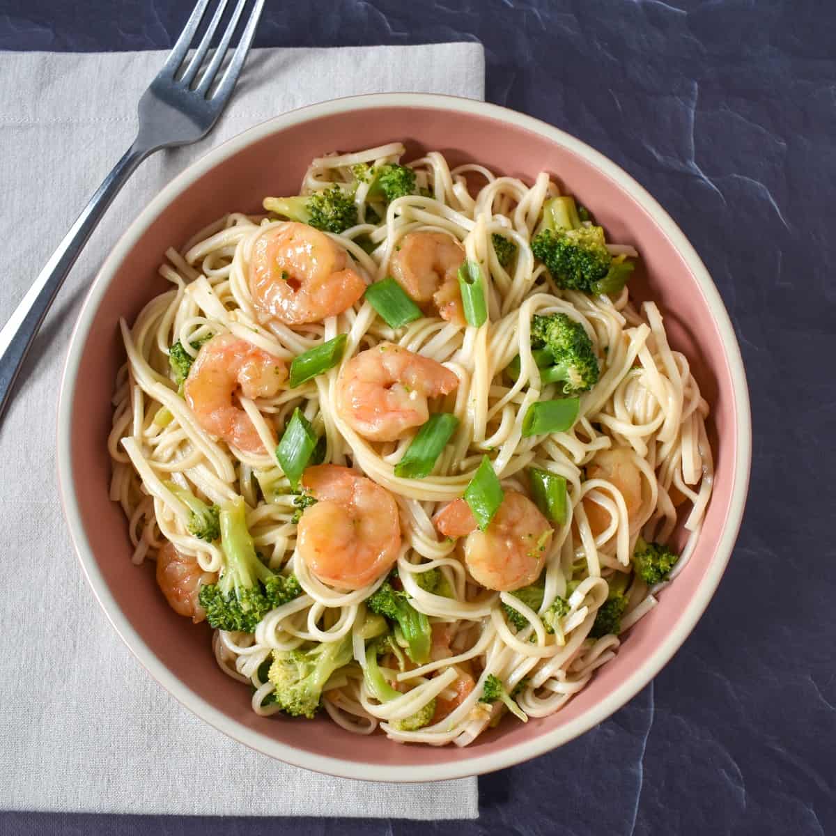 An image of the shrimp lo mein served in pink plate and set on a beige linen on a black table.