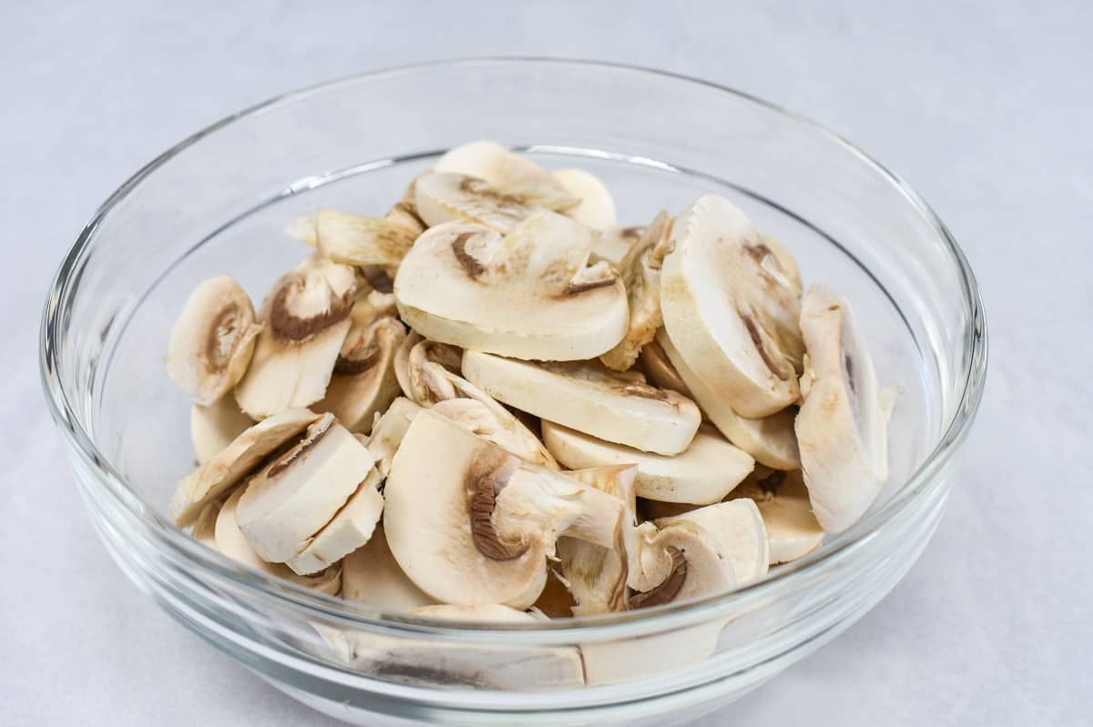An image of sliced mushrooms in a glass bowl, set on a white table.