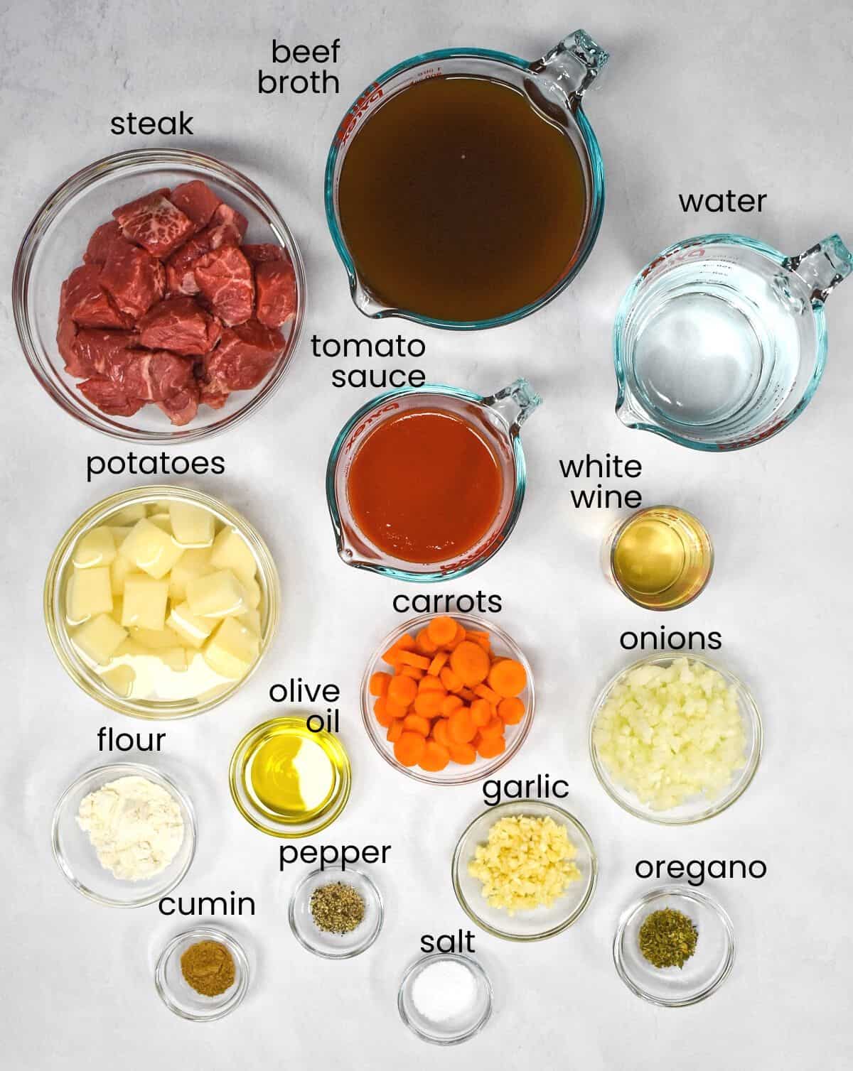 The ingredients for the beef soup arranged in glass bowls on a white table with each ingredient labeled with small, black letters.