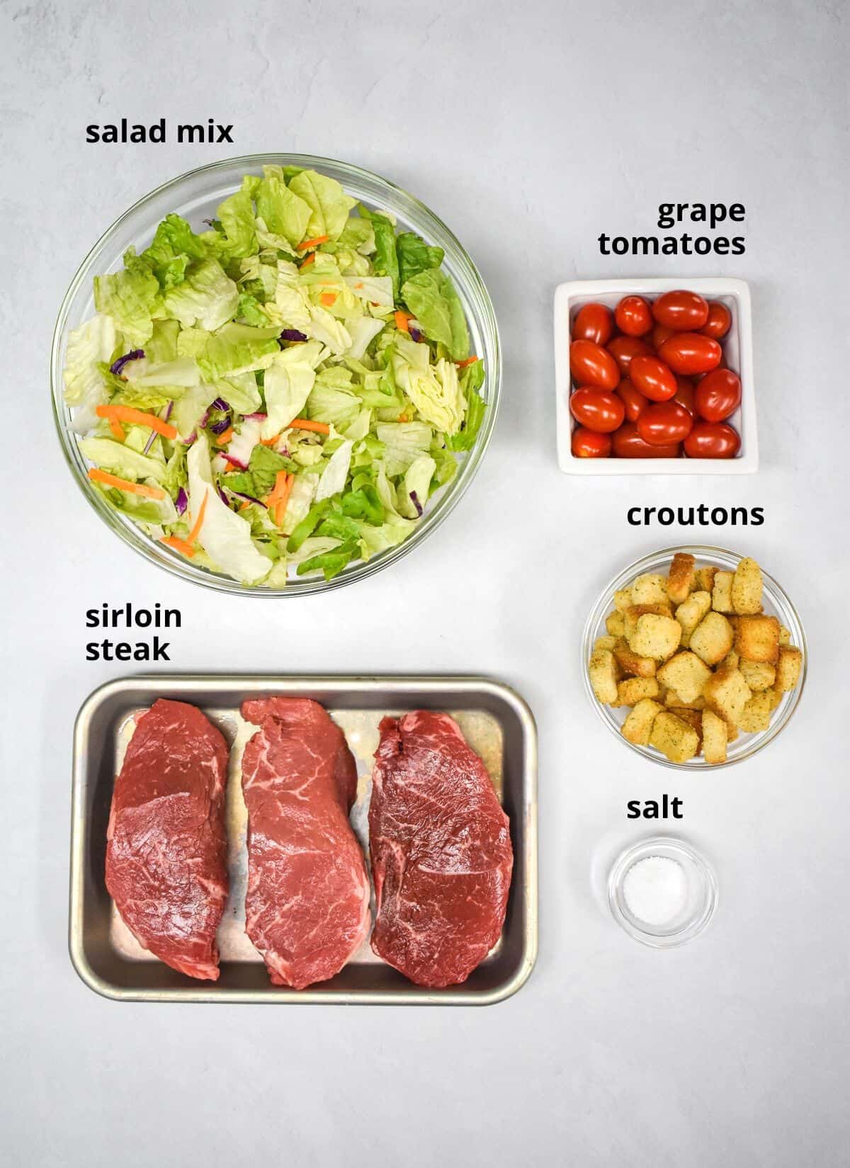 The ingredients for the steak salad arranged on a white table with each ingredient labeled in black letters.