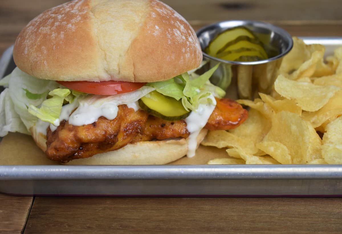Buffalo Chicken Sandwich served with lettuce, tomato, pickles, and blue cheese dressing on a bun with a side of chips